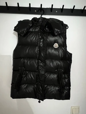 Svart dunväst från Moncler - Snygg svart dunväst från Moncler med huva och två dragkedjefickor framtill. Västen har Moncler-logga på bröstet och quiltad design som ger en puffig look. Perfekt för lager-på-lager och streetstyle. Materialet är glansigt och känns lyxigt.