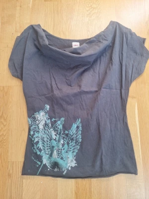 Grå topp med turkost tryck från ONLY - Säljer en grå topp från ONLY med lös passform och korta ärmar. Toppen har ett stort tryck i turkost och glitter  med vingar och gitarr på framsidan samt en bred, avslappnad halsringning. Perfekt för dig som gillar coola detaljer och unik design.