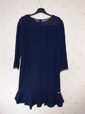 Marinblå glittrig klänning från Aldona - Snygg marinblå klänning från Aldona med glittrigt tyg, volangkant nertill och dekorativ axeldetalj. Klänningen har trekvartsärm och meshparti upptill som ger en elegant touch. Perfekt för dig som vill sticka ut lite extra.