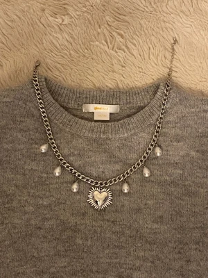Silverfärgat halsband med pärlor och hjärta - Snyggt silverfärgat halsband från Gina Tricot med chunky kedja, dekorativa vita pärlor och ett stort hjärthänge med solstråle-design. Perfekt statement piece för att lyfta din outfit och ge en cool touch.