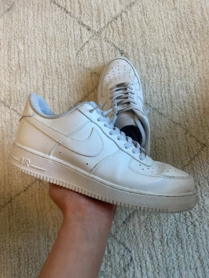 Vita Nike Air Force 1 sneakers - Klassiska vita Nike Air Force 1 sneakers i lågt utförande. Skorna har en stilren design med perforerad tå, vit sula och ikoniska swoosh-loggan på sidan. Tillverkade i läder med textilfoder och snörning framtill. Perfekta för dig som gillar tidlös streetstyle.