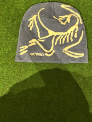 Blå mössa från Arc'teryx - Snygg grå mössa från Arc'teryx med ikoniskt fossilmönster i ljusgul färg på framsidan. Mössan är stickad och har en klassisk, tight passform. Perfekt för kalla dagar när du vill hålla stilen och värmen.