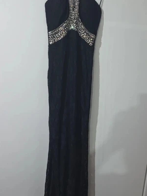 Svart långklänning med paljettdetaljer - Snygg svart långklänning med glittriga paljettdetaljer över bysten och axelbanden. Klänningen har en figurnära passform och är ärmlös med en elegant öppen rygg. Perfekt för dig som vill sticka ut på bal eller fest.