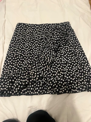 Svart kortkjol med vita blommor - Säljer en svart kortkjol med små vita blommor och volangdetalj. Kjolen har omlottkänsla och är perfekt för dig som gillar en söt och trendig stil. Passar till många olika toppar och sneakers.