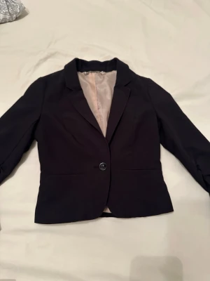 Svart klassisk kavaj med knapp - Stilren svart kavaj med en knapp framtill och ljus innerfoder. Kavajen har klassisk krage, två fickor och figurnära passform. Perfekt för en snygg och enkel look. Materialet känns mjukt och följsamt.