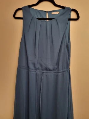 Blå långklänning med v-ringning - Petrolblå ärmlös långklänning med under klänning. Klänningen har en enkel och stilren look med markerad midja och ett mjukt, flytande tyg som ger ett elegant fall. Delvis öppen i ryggen.