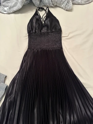 Svart långklänning i satin - Elegant svart långklänning med halterneck och smockad midja. Klänningen har plisserad kjol och djup v-ringning, tillverkad i glansigt satinliknande material. Perfekt för dig som vill ha en stilren och chic look.