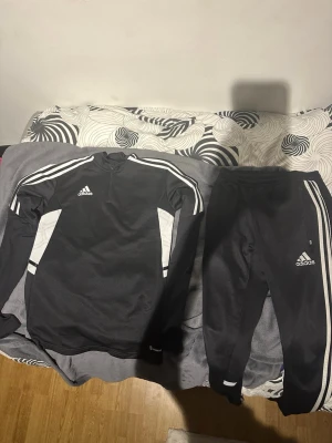 Svart Adidas träningsset med vita detaljer, använda en gång och tvättade - Säljer ett svart träningsset från Adidas med vita detaljer. Setet består av en långärmad tröja med halv dragkedja och matchande träningsbyxor med dragkedja vid fickorna. Båda plaggen har klassiska Adidas-ränder och logga på bröst och ben. 