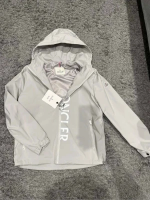 Ljusgrå vindjacka från Moncler - Snygg ljusgrå vindjacka från Moncler med stor logga framtill. Jackan har huva, dragkedja, två fickor med dragkedja och justerbar nederkant. Tillverkad i ett lätt och vattenavvisande material, perfekt för blåsiga dagar. Moncler-märke på ärmen för extra stilpoäng.