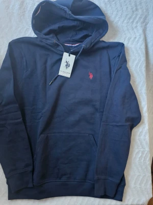 Marinblå hoodie från U.S. Polo Assn. - Snygg marinblå hoodie från U.S. Polo Assn. med klassisk huva och dragsko. Tröjan har en röd broderad logga på bröstet och en stor magficka framtill. Tillverkad i mjukt bomullsmaterial som känns skönt mot huden. Perfekt för en avslappnad och stilren look.