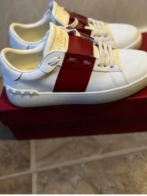 Valentino open - Snygga vita sneakers från Valentino med bred röd läderdetalj över mitten och nitar på hälen. Klassisk låg modell med vita snören och gummisula. Märkeslogga på plösen och undersidan. Perfekta för dig som gillar exklusiv streetstyle.
