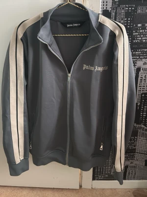 Grå ziptröja från Palm Angels - Snygg grå ziptröja från Palm Angels med vita och svarta ränder längs ärmarna. Tröjan har hög krage, dragkedja framtill och två fickor med dragkedja. Palm Angels-logga broderad på bröstet. Perfekt för en sportig och trendig look.