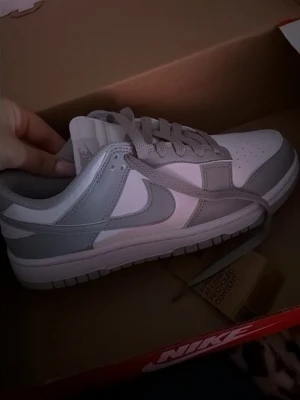 Nike Dunk Low grå/vit sneakers - Nike Dunk Low sneakers i stilren grå och vit färgkombination. Skorna har klassisk låg siluett, perforerad tåbox och ikoniska swoosh-loggan på sidan. Ovandelen är i läder med matchande grå snören och vit mellansula. Perfekta för en clean och trendig look.