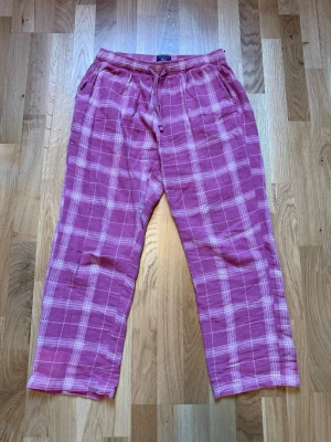 Rosa rutiga pyjamasbyxor - Mysiga pyjamasbyxor i rosa med vitt rutmönster. Byxorna har resår i midjan och dragsko för extra komfort samt sidofickor. Perfekta för chillkvällar hemma. Materialet känns mjukt och luftigt.