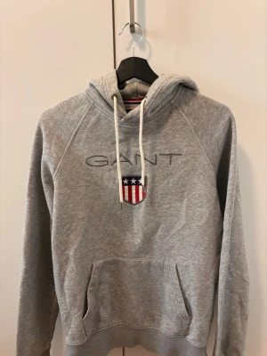 Grå hoodie från GANT med logga - Snygg grå hoodie från GANT med broderad logga och amerikanskinspirerat sköldmärke på bröstet. Hoodien har vit dragsko i huvan och en stor magficka. Tillverkad i mjukt sweatshirtmaterial, perfekt för en avslappnad stil.