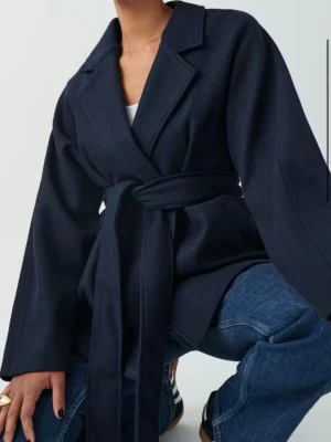 Mörkblå kappa med knytskärp - Stilren mörkblå kappa med breda slag och knytskärp i midjan. Kappan har en rak passform och är tillverkad i ett mjukt tyg som ger en snygg siluett. Perfekt att slänga över en hoodie eller tröja för en chill look.