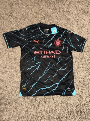Manchester City bortatröja Puma - Snygg oanvänd Manchester City tröja från Puma i svart med turkosa blixtmönster och rosa detaljer. Tröjan har klubbmärke och Puma-logga på bröstet samt Etihad Airways som sponsortryck. Tillverkad i ett lätt och funktionellt material.