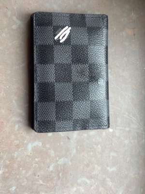 Svart plånbok med rutmönster - Snygg svart plånbok L. V.  med klassiskt rutmönster och flera kortfack. Tillverkad i slitstarkt konstläder med stilren design. Perfekt för dig som vill ha en smidig och trendig accessoar till dina kort och kontanter.