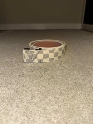 Damier-bälte beige - Snyggt beige bälte  med det klassiska Damier-mönstret och en silverfärgad logga som spänne. Bältet är gjort i läder och har en stilren, modern look som passar till jeans eller byxor. Perfekt accessoar för dig som gillar exklusiva detaljer.