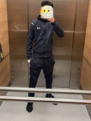 Svart träningsset från Under Armour - Säljer ett svart träningsset från Under Armour med zip hoodie och matchande byxor. Setet har vita detaljer och logga på både överdel och underdel. Perfekt för gymmet eller sportaktiviteter. Materialet är mjukt och stretchigt för maximal komfort.