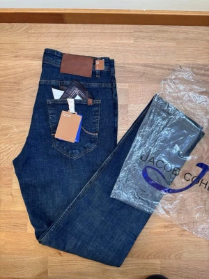Mörkblå jeans från Jacob Cohën - Snygga mörkblå jeans från Jacob Cohën med klassisk femficksmodell och bruna kontrastsömmar. Jeansen har en patch i läder bak i midjan och detaljerad innerficka med mönster. Tillverkade i mjuk denim med exklusiv känsla. Perfekta för dig som gillar stilrena och tidlösa jeans. Skicka gärna bud och skriv vid minsta fråga 