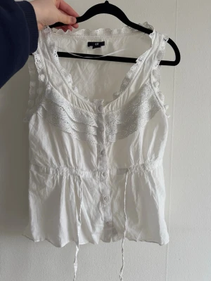 Vit spetsdetaljerad linnetopp H&M - Söt vit linnetopp från H&M med spetsdetaljer längs axelband och urringning. Toppen har knappar framtill, justerbar midja med knytband och är ärmlös. Perfekt för en fräsch och somrig look.