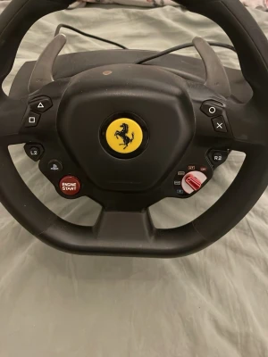 Thrustmaster Ferrari ratt till PS4/PC - Säljer en svart Thrustmaster ratt inspirerad av Ferrari, perfekt för racing på PS4 eller PC. Ratten har flera knappar, paddlar för växling och en tydlig Ferrari-logga i mitten. Materialet är mestadels plast och designen är sportig och robust.