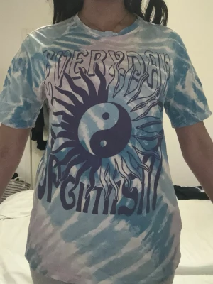Tie-dye t-shirt med tryck - Säljer en blå, lila och vit tie-dye t-shirt med stort grafiskt tryck framtill som visar en sol och yin-yang-symbol samt texten 'EVERYDAY OPTIMISM'. T-shirten har korta ärmar och en relaxed fit. Perfekt för dig som gillar unika och färgglada plagg. Storlek S men passar mig som är M.
