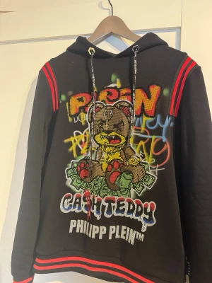 Philipp Plein Cash Teddy hoodie - Svart hoodie från Philipp Plein med färgglad graffiti-inspirerad björn och texten 'CASH TEDDY' framtill. Hoodien har röda detaljer på axlar och nederkant, samt dragsko i huvan. Tillverkad i 100% bomull för skön känsla och streetwear-vibe.