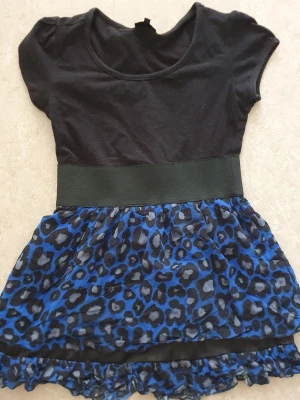 Klänning - Kort klänning från KappAhl med svart överdel och korta ärmar. Kjoldelen har blått och svart leopardmönster samt volangkant nertill. Bred midjeresår för skön passform. Perfekt för dig som gillar djurmönster och vill sticka ut.