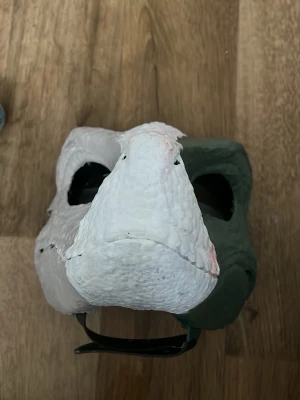 Halvmask dinosaurie cosplay - Cool halvmask i hårdplast som föreställer en dinosaurie med vit och grön färg. Masken har tydliga näsborrar och struktur som liknar reptilskinn. Perfekt för maskerad eller cosplay och sitter med ett elastiskt band runt huvudet.