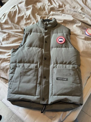 Grön dunväst från Canada Goose - Snygg grå dunväst från Canada Goose med hög krage och två stora framfickor. Västen har det klassiska Canada Goose-märket på bröstet och stängs med både dragkedja och knappar. Perfekt för lager-på-lager och håller dig varm under kyliga dagar. Helt ny nästan storlek m
