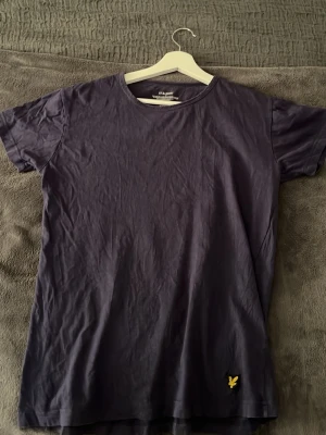Mörkblå Lyle & scott T-shirt - En stilren mörkblå t-shirt med rund hals och korta ärmar. På framsidan finns en liten gul broderad logga nertill. Perfekt för en avslappnad och enkel look.