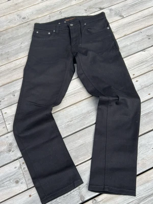 Svarta raka jeans från Nudie Jeans - Snygga svarta jeans från Nudie Jeans med klassisk femficksmodell och raka ben. Jeansen har silverfärgade nitar och knapp, samt diskret logga på insidan av linningen. Tillverkade i ett slitstarkt denimtyg som känns både trendigt och tidlöst.