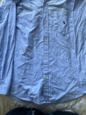 Ljusblå skjorta från Polo Ralph Lauren - Klassisk ljusblå skjorta från Polo Ralph Lauren med knappslå och bröstficka med broderad logga. Skjortan har lång ärm, button-down krage och är tillverkad i mjuk bomull. Perfekt för en clean och stilren look.