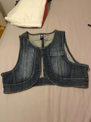 Jeans bolero - Säljer en croppad jeansbolero i blå denim med markerade sömmar och rundad nederkant. Västen har en fickdetalj framtill och är ärmlös, perfekt för att addera en cool touch till din outfit.