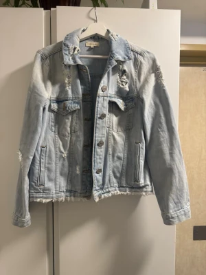 Ljusblå jeansjacka River Island - Trendig ljusblå jeansjacka från River Island med slitna detaljer och råa kanter. Klassisk modell med bröstfickor med lock, knappar framtill och raka ärmar. Perfekt för en avslappnad och cool stil.