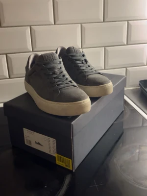 Gråa sneakers från Marc O'Polo - Snygga grå sneakers i mocka från Marc O'Polo med vit sula och klassisk låg profil. Skorna har rund tå, matchande grå skosnören och diskret logga på plösen. Perfekta för en stilren och avslappnad look.