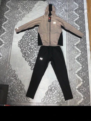 Under Armour  - Säljer ett snyggt träningsset från Under Armour med beige och svart jacka med huva och svarta byxor. Jackan har dragkedja, reflekterande logga och svarta paneler på sidorna. Byxorna är svarta med resår i midjan och reflekterande detaljer vid benen.