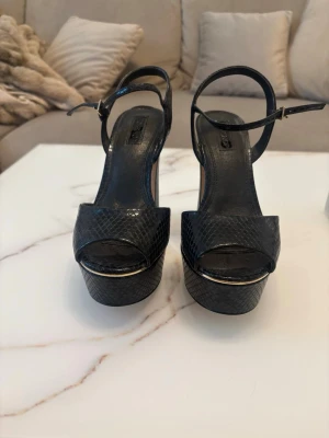 Svarta platåsandaler med ormmönster - Säljer ett par svarta sandaler med hög platå och bred klack. Skorna har ett snyggt ormmönster och justerbart ankelband med spänne i guld. Öppen tå och stilren design som ger extra höjd och attityd till din outfit.