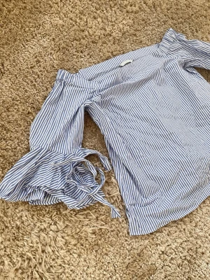 Blåvit offshoulder blus med volangärm - Snygg blus med blå och vita ränder i offshoulder-modell. Långa ärmar med volang och knytband vid ärmslut ger en trendig look. Perfekt till jeans eller kjol för en fräsch stil. Tunn och luftig bomullskänsla.