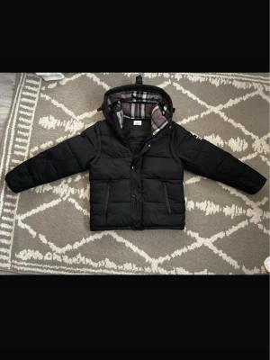 Burberry jacka - Säljer en svart pufferjacka med huva och rutigt foder i grått, vitt och rött. Jackan har två framfickor med dragkedja och tryckknappar framtill. Perfekt för kalla vinterdagar och har en modern, boxig passform.