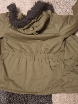 Olivgrön parkas med fuskpäls - Snygg olivgrön parkas med huva och svart fuskpäls. Jackan har dragsko i midjan för en schysst passform och stora fickor framtill. Insidan är fodrad med mjukt material som håller dig varm under kalla dagar. Perfekt för dig som vill ha en stilren och praktisk jacka.