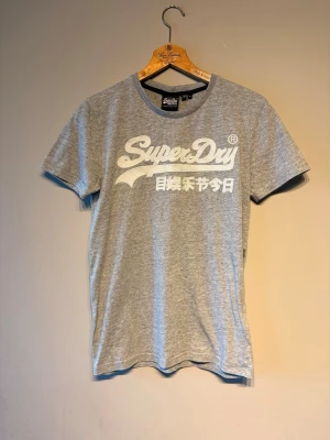 Grå Superdry t-shirt med tryck - Snygg grå t-shirt från Superdry med stort vitt tryck på bröstet och japanska tecken. Klassisk rund halsringning och korta ärmar. Perfekt till jeans eller shorts för en avslappnad stil.