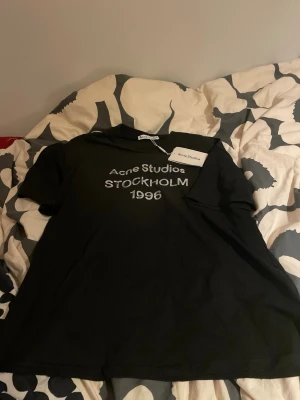 Svart Acne Studios t-shirt - Svart t-shirt från Acne Studios med tryck 'Acne Studios STOCKHOLM 1996' på bröstet. Klassisk passform med rund hals och korta ärmar. Perfekt för en stilren och trendig look. Köpt på Vinted för dubbla priset men har aldrig kommit till användning 