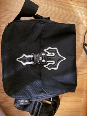 Svart axelväska från Trapstar - Säljer en svart axelväska från Trapstar med justerbar rem och coolt metallspänne framtill. Väskan har en mix av slätt och mönstrat material samt Trapstar-logga på remmen. Perfekt för att bära det viktigaste med stil.