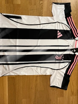 Juventus matchtröja - Juventus matchtröja från Adidas i svartvitt med rosa detaljer och tre rosa ränder på axlarna. Tröjan har klubbmärke och Adidas-logga på bröstet, rund hals och är tillverkad i ett lätt, ventilerande material perfekt för träning eller match.   Fraktar samma eler dagen efter du köpt!!! Endast en kvar i lager!!!