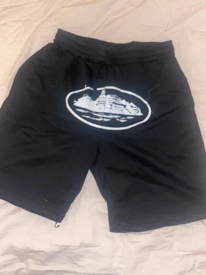 Svarta shorts med tryck - Svarta shorts med resårmidja och snörning. På framsidan finns ett stort vitt tryck av ett fartyg i en oval. Shortsen har en loose passform och är perfekta för sommaren. Materialet känns som bomull eller polyester.