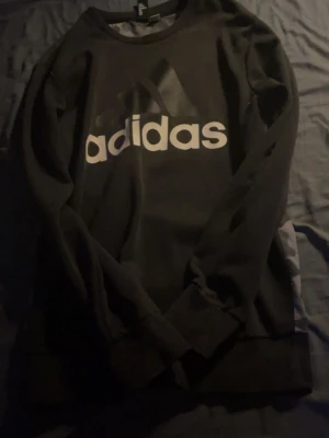 Svart Adidas sweatshirt med camomönster - Säljer en svart sweatshirt från Adidas med stort vitt logotryck framtill och camomönstrad rygg. Tröjan har rund halsringning och långa ärmar med ribbade muddar. Perfekt för dig som gillar streetwear och sportig stil.