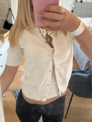 Vit kortärmad kofta med knappar - Snygg vit kofta med korta ärmar och knäppning framtill. Klassisk rund halsringning och ribbade muddar vid ärmslut och nederkant. Perfekt att styla över ett linne eller t-shirt för en clean och fräsch look.